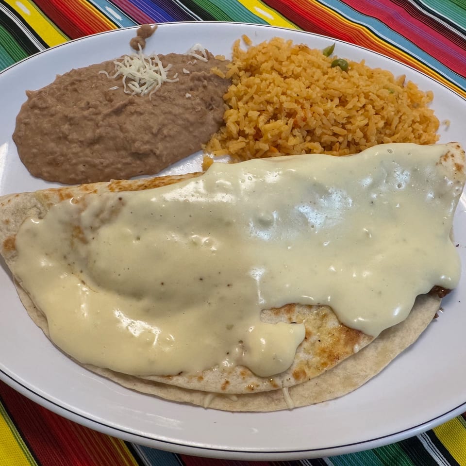 Cheese-covered burrito served with refried beans and Mexican rice on a white plate over a colorful striped tablecloth.