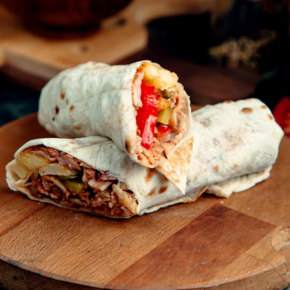Two Mexican burritos, one cut in half, filled with meat, red peppers, and potatoes on a wooden board.