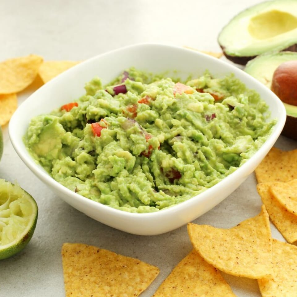 Fresh guacamole in a white bowl, surrounded by tortilla chips, lime, and avocado halves.